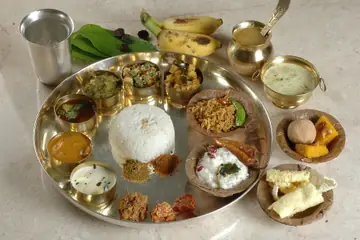 A steel plate of rice with small bowls of curries, curd, pickle and sweets, with bananas and a glass of water beside it