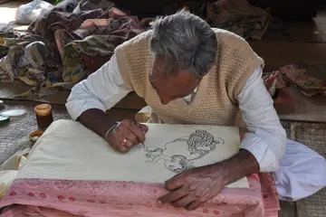 An artist bending over a cloth and drawing a Kalamkari outline in ink
