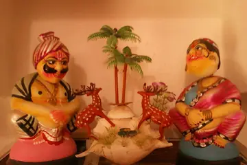 A pair of painted Kondapalli toy figures, a man in a turban and a woman in a saree, with two red deer and a small palm between them