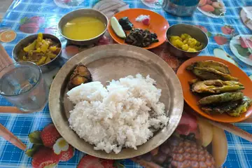 A brass plate of rice with bowls of dal, vegetables, greens and fried fish on a table