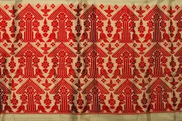 A cream gamosa laid flat, its red woven flowers, birds and lamps in rows between striped red ends