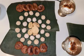 Pitha, round white coconut laru and brown ghila pitha laid in rings on a banana leaf, with two small cups of curd and jaggery
