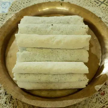 A stack of white rice-flour til pitha rolls on a round brass plate