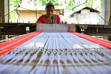 A woman at a loom in a wooden shed with white warp threads stretched in front of her and red bands at each side