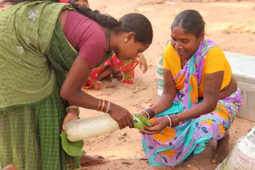 Two women in colourful saris crouched at a weekly haat in Bastar, one holding a bottle and the other a leaf
