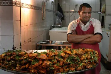 A cook in an apron standing with arms folded behind a big tray of fried food in a kitchen in Chandigarh