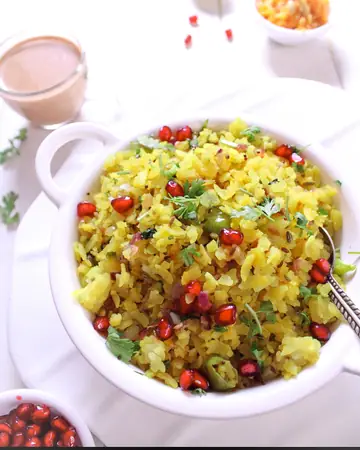 A white bowl of yellow kanda poha topped with pomegranate seeds, green chillies and coriander