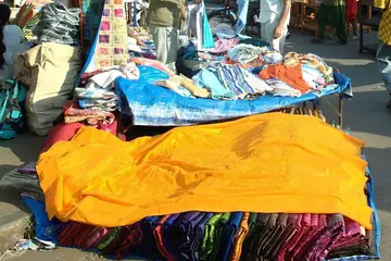 Bundles of printed and plain cloth, one bright orange, heaped on blue sheets at a street market in Mapusa, Goa