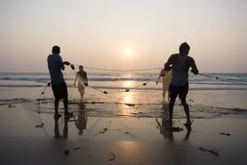 Two men in silhouette hauling a fishing net in the shallows at sunset at Arambol, Goa