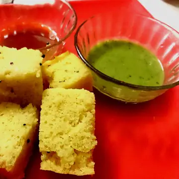Squares of yellow dhokla with mustard seeds and a bowl of green chutney.