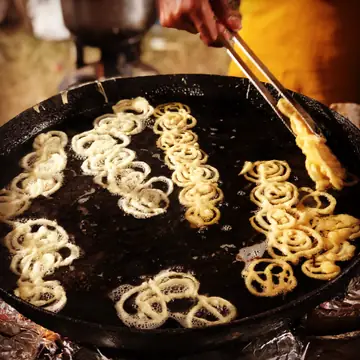 Jalebis frying in a wide black pan while a hand turns them with tongs