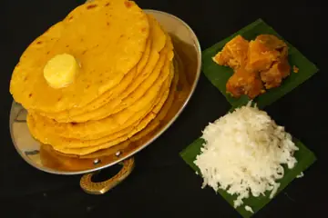 A stack of golden makki rotis with butter, beside jaggery and grated radish on leaves