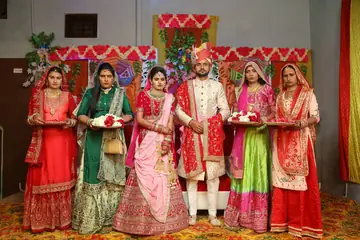 A bride, a groom and four women in red, green and pink wedding clothes holding flower plates, in Haryana