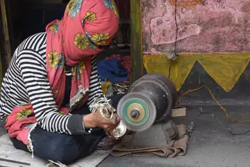 छपे हुए स्कार्फ़ में एक कारीगर औरत पॉलिश के पहिये पर पीतल की छोटी मूर्तियाँ चमकाते हुए