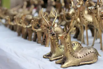 ढोकरा धातु शिल्प के दो लेटे हुए पीतल के हिरण, जिन पर बारीक तार की नक़्क़ाशी है, पीछे धुँधले और हिरण