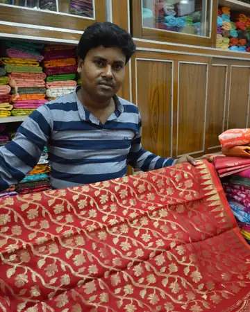 Saree ki dukaan mein ek seller sunhare phoolon wali laal tussar saree ko haath mein pakde dikha raha hai