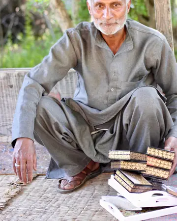 A craftsman sits on a mat beside a stack of painted papier-mâché boxes.