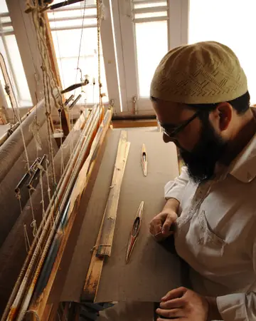 A weaver in a white cap working at a wooden hand loom beside a window in Srinagar