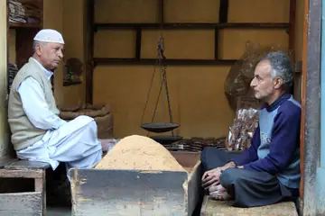 Two shopkeepers sitting in a small shop beside a hanging scale and a heap of flour