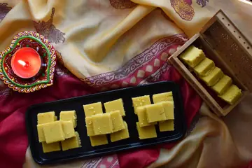 Squares of yellow Mysore Pak sweet on a black tray beside a lit diya