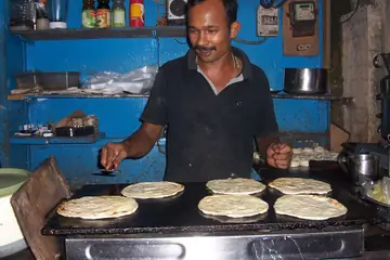 A cook in Kochi turning flat parottas on a griddle at his stall, with bottles on the shelves behind him