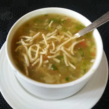 A white bowl of vegetable soup topped with fried noodles and a steel spoon