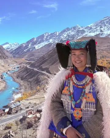 A smiling woman in a dark perak headdress, a blue embroidered coat and a woollen fur-edged cloak, a river valley behind her