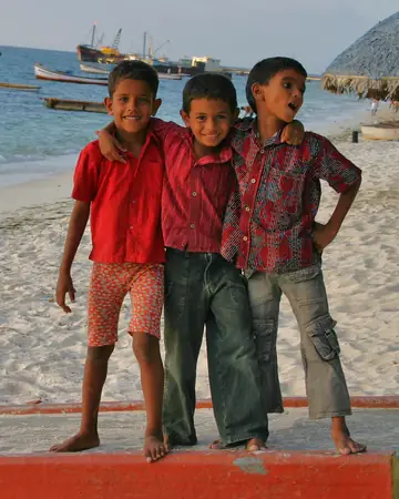 Three smiling children in red shirts standing arm in arm on a beach at Kavaratti, boats in the lagoon behind