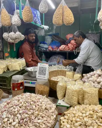 Two men at a stall in Crawford Market, Mumbai, with heaps of garlic, bags of puffed snacks and a weighing scale on the counter