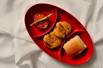 A fried batata vada and a pav with red chutney and a green chilli on a red oval tray