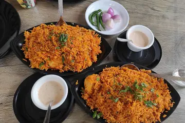 Two black plates of orange chicken biryani with cups of milk tea on a wooden table