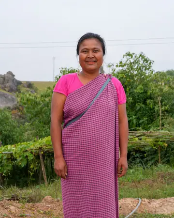 A woman in a pink checked wrap and pink blouse standing by a field in Mawphanlur