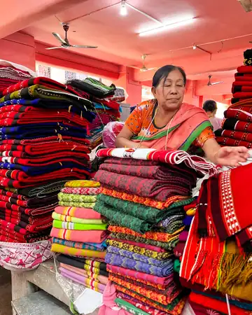 A woman sitting among tall stacks of woven shawls and cloth in a market hall in Imphal