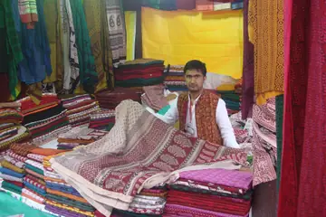 Bagh print ka kaarigar apne thele par, jahan chhape hue kapde ke dher lage hain aur ek chhapa hua bedcover ooncha pakda hai