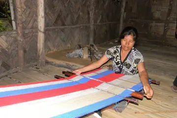 A woman weaving red, white and blue striped cloth on a loom in a bamboo-walled house