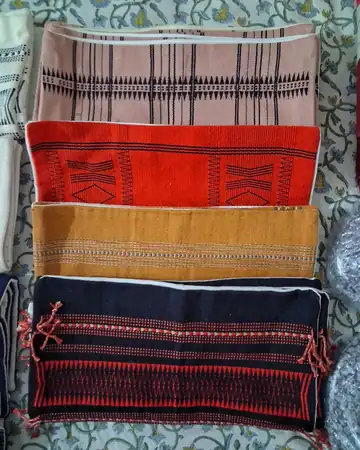 Four folded hand-woven shawls in cream, red, gold and dark green, stacked in a pile