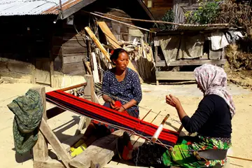 Two women at a wooden loom weaving a red striped cloth in a sunny yard