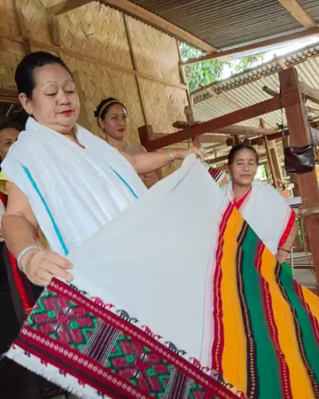 Women in a weaving shed holding up a white shawl with a red patterned border