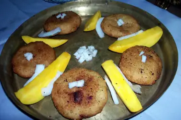 Round fried kakara pithas on a steel plate with slices of yellow mango and bits of coconut