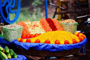 A round basket heaped with orange and yellow snack mix, ringed with tomatoes, with cucumbers beside it on a blue cloth
