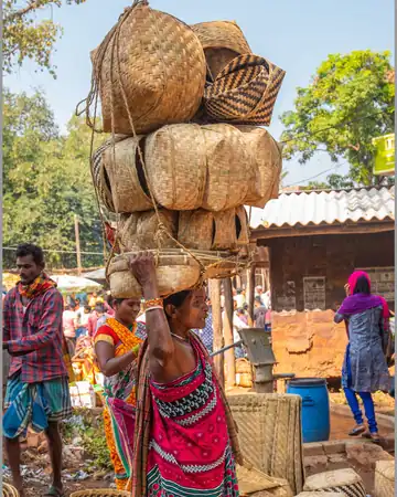 A woman balancing a tall stack of cane baskets on her head at the weekly haat in Doraguda