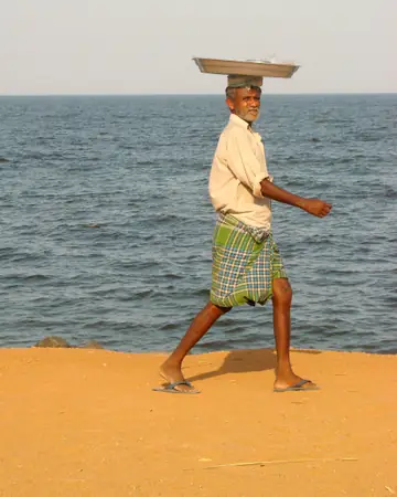 A man in a checked lungi walking along the sand with a flat tray on his head, the sea behind him