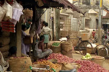 Sacks of red onions and potatoes in a covered market lane, a seller sitting among them and bicycles parked beside