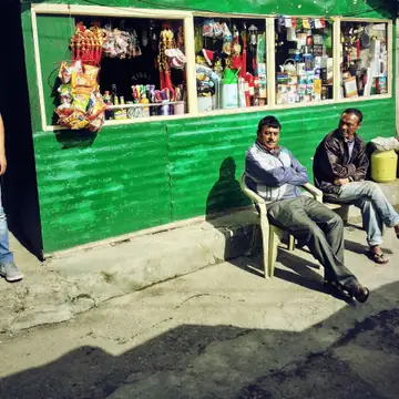 A green wooden hill shop kiosk with goods in its window and two men sitting on chairs in front