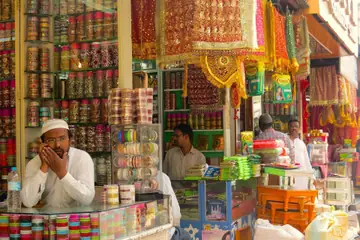 A shopkeeper resting his chin on his hand behind a glass counter, with shelves of bangles behind him in Laad Bazaar, Hyderabad