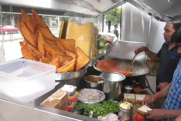 Cooks frying and assembling snacks at a street food stall, fried snacks piled at the left