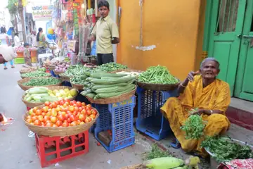 A woman in a yellow saree sitting among baskets of tomatoes, beans and gourds on a street in Hyderabad, a man standing behind