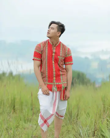 A man in a red woven shirt and a white dhoti standing in tall grass with hills behind