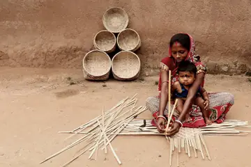 A woman weaving thin bamboo strips on the ground beside a stack of round bamboo baskets, a baby on her lap