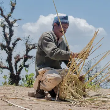 A man in a cap crouched on a hilltop weaving ringal bamboo strips for a basket against a blue sky